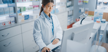 "Pharmacist Pharmacist standing in front of a computer looks at the screen as she moves the computer’s mouse.